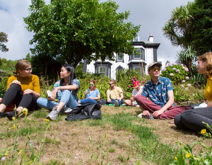 Students sat on the grass at Falmouth campus in front of an old black and shite Victorian building.