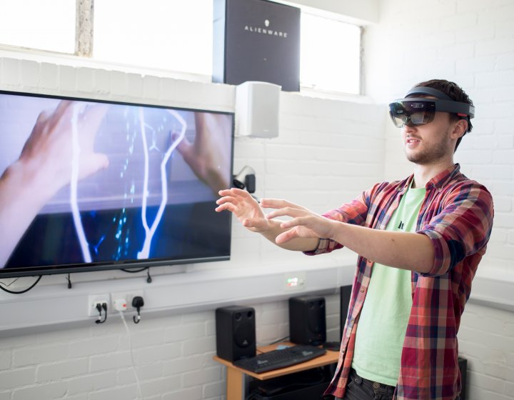 Student wearing virtual reality headset with arms extended in front and images of hands and electricity bolts on screen next to him.