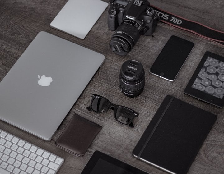 A desk with laptop, keyboard, camera, lens, smartphone.