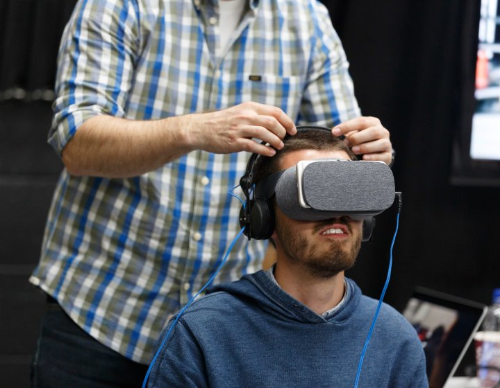 man seated wearing VR headset with another man stood behind him