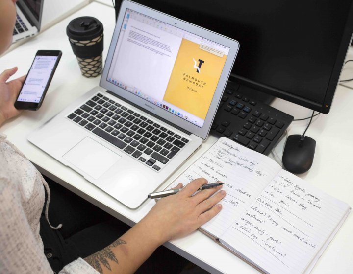 A Journalism student working at a laptop and holding a phone