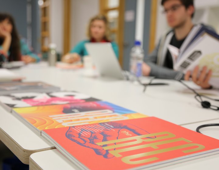 Design journals on a desk in the foreground, students working out of focus in the background.