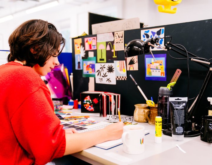Falmouth University Illustration student wearing a red jumper and drawing at a desk