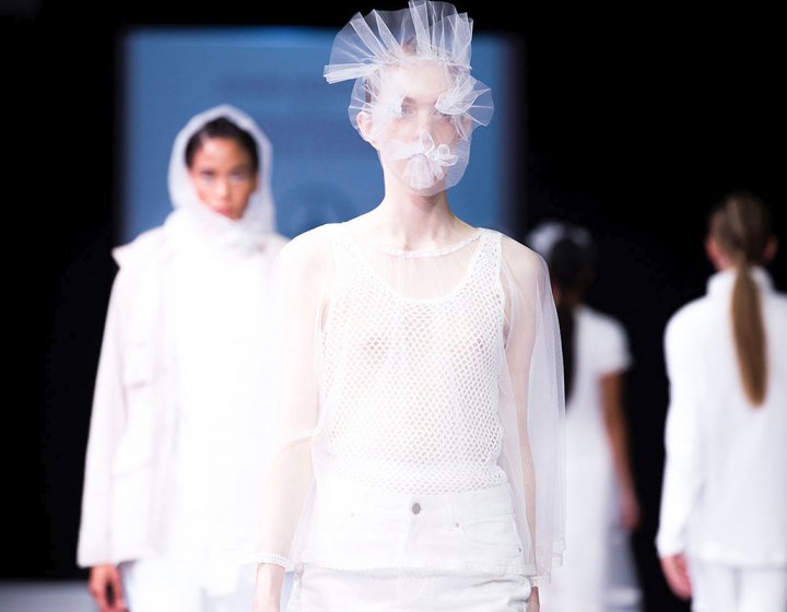 Models on a catwalk wearing white clothing and a netted face mask
