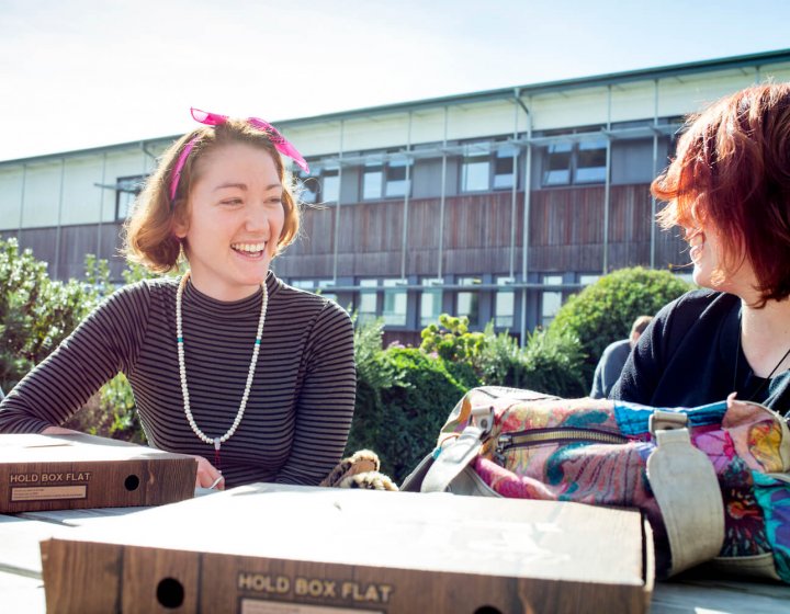 Two female students smiling sat at a picnic bench on Penryn Campus.