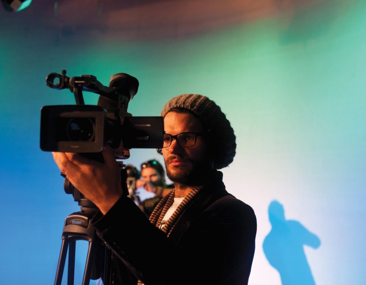 A male student wearing a hat and operating a film camera in a blue studio