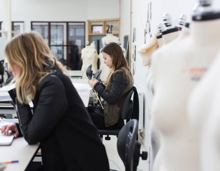 Two students in fashion studio, one on laptop and one knitting.