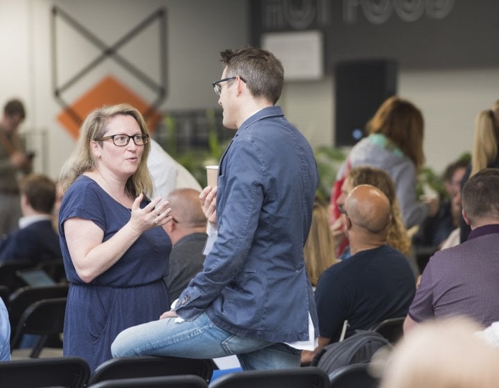 A man and a woman standing and talking in a room full of people