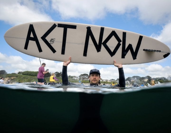 Activist in the sea holding up a surfboard with Act Now message written on it
