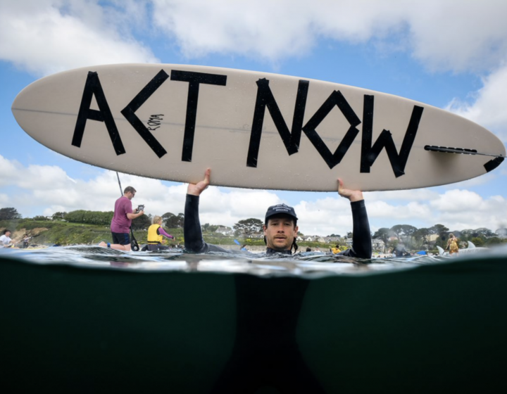 Activist in the sea holding up a surfboard with Act Now message written on it