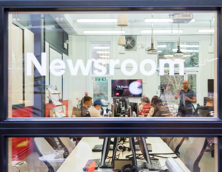 Through the window of the newsroom with lecturer talking to journalism students.