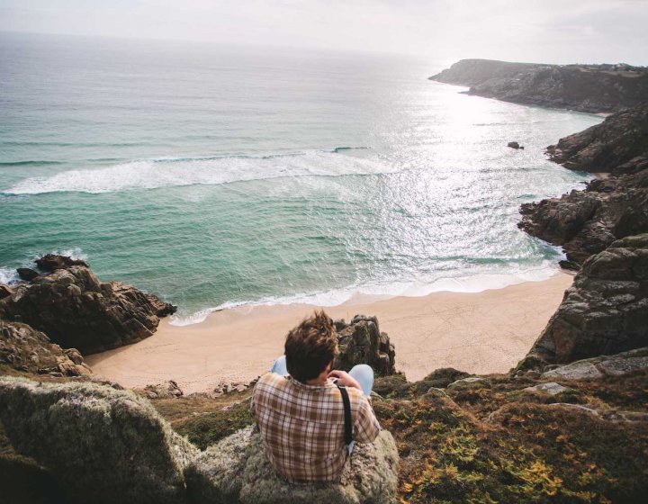 Man sat on the edge of cliff looking out to sun reflections on the water and coastline.