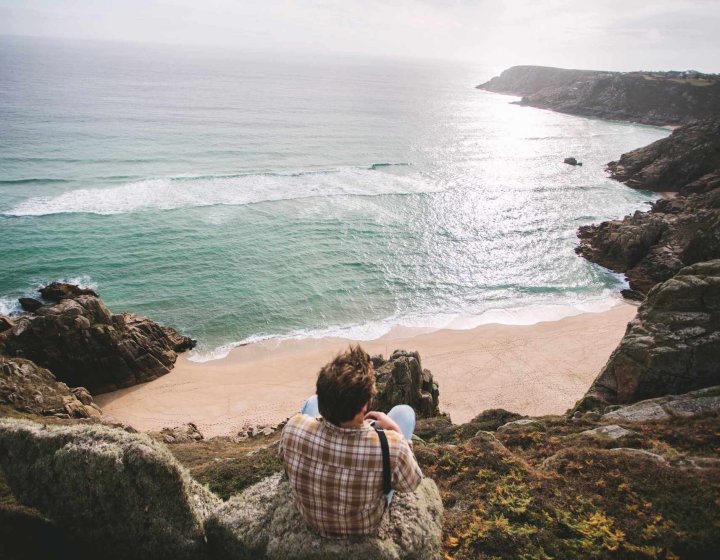 A Falmouth University student sitting on a cliff top overlooking a beach and sea
