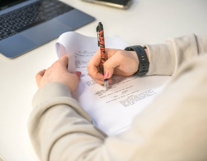 a person holding a pen and annotating a script with a laptop in the background