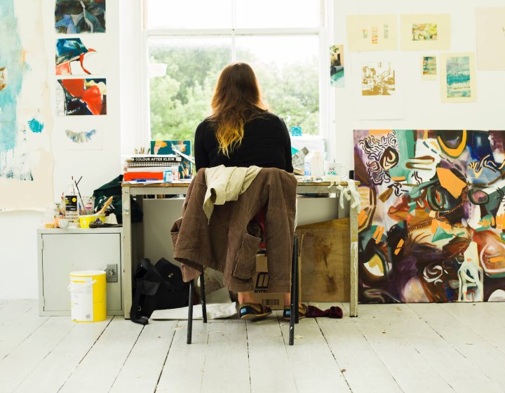 Back of student sitting at desk in front of window surrounded by artwork and paintings.