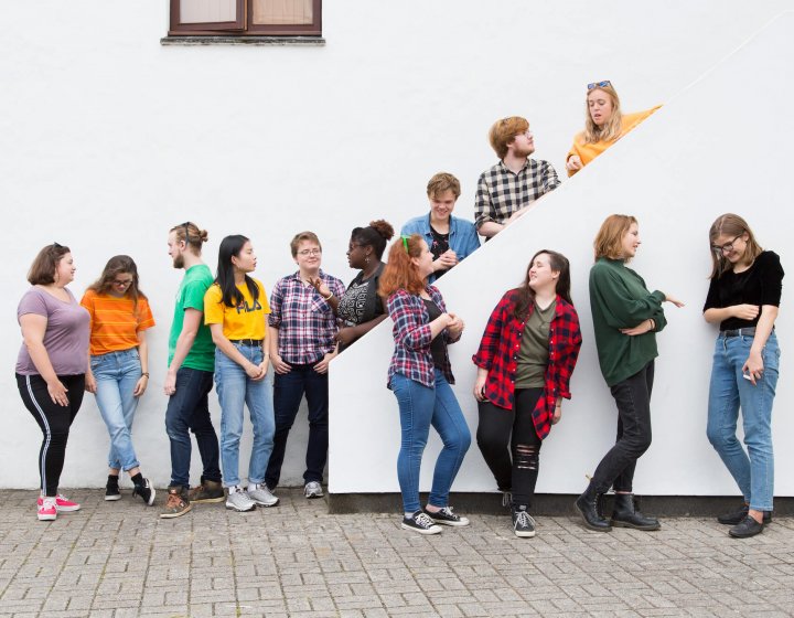 13 Falmouth University students in different coloured tops stood against a white wall and on a white staircase.