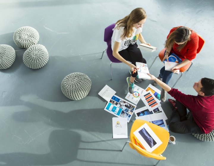 Falmouth University Business students sat on chairs on grey floor looking at work together.