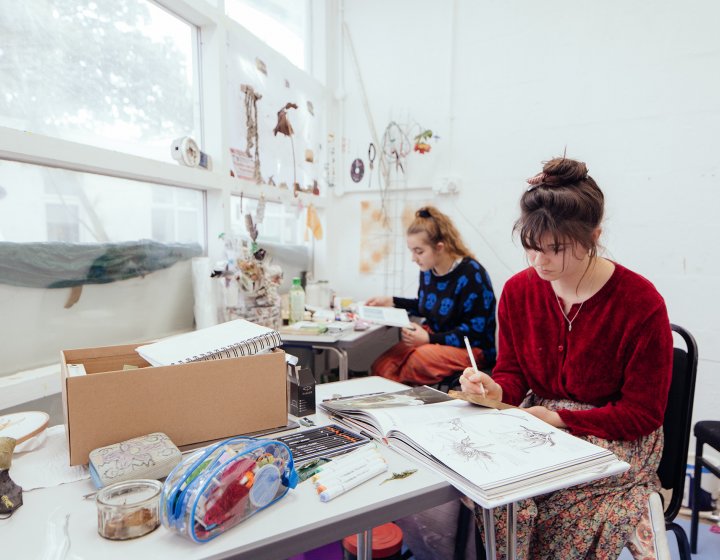 Two art students sat at desks in front of large window and drawing.