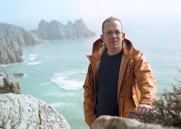 Writer, Wyl Menmuir, standing on cliff with sea view in the background.
