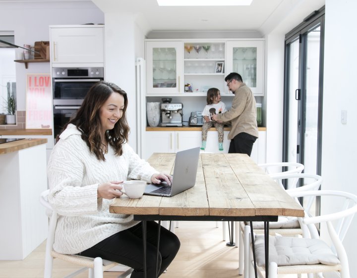 Woman sits at her dining room table with a laptop whilst her husband and child are in the background.
