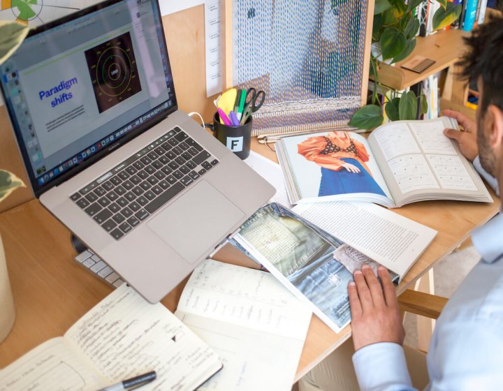 A man sat at a computer with fashion-related books and paper