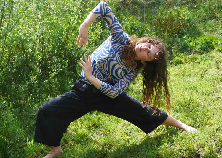 A person dancing outside on green grass with a bush in the background
