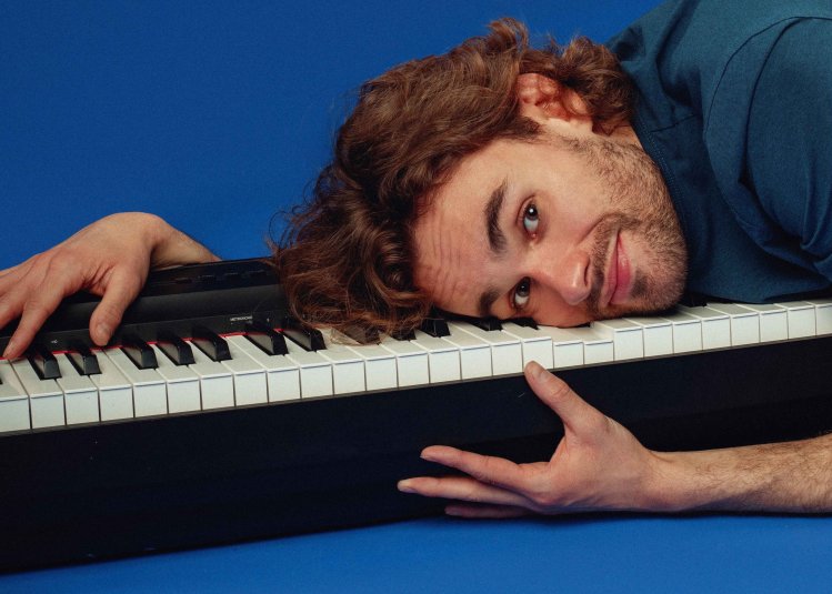 A person laying on top of a piano keyboard against a royal blue backdrop