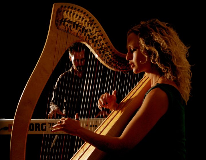 A close image of a harpist holding and playing the instrument with a person in the background playing an electric piano