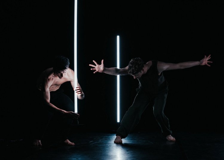 Two dancers on stage in very dim light performing around two white strip lights