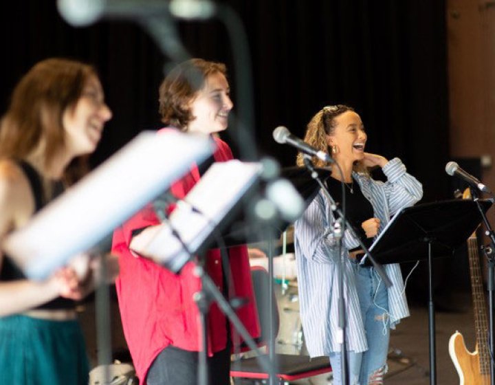 Three performers holding sheet music singing into microphones on stage