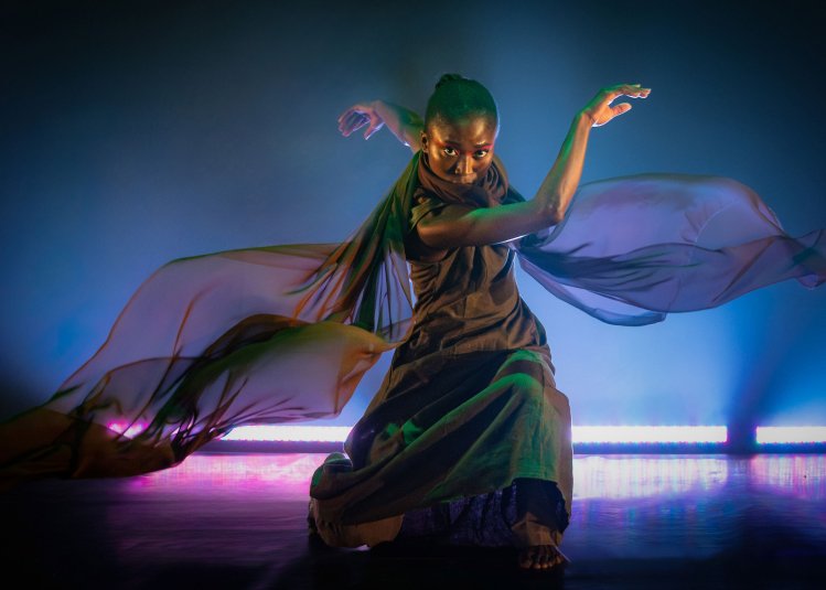 A dancer wearer colourful costume with a pink/blue uplighter behind them