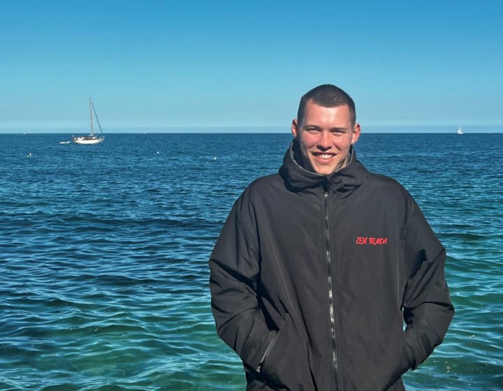 Portrait shot of Ben Penrose wearing black beach robe with red Zen Beach logo, with blue sea in the background