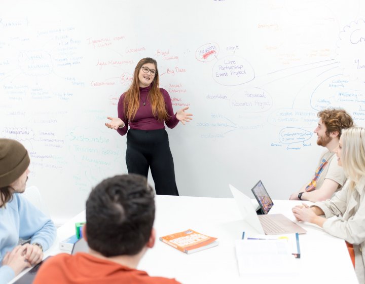 A student leads a discussion, with a large whiteboard full of notes behind her.