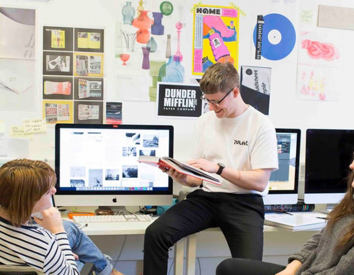 Three Falmouth University students sat round an iMac with graphic design posters on the wall