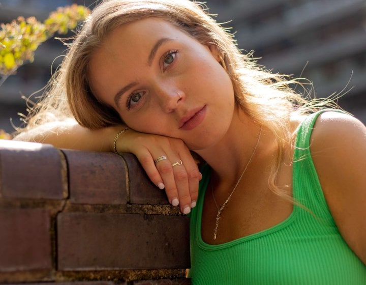 Graduate Eva Gadd wearing a bright green tank top and leaning against a red brick wall, with large block of flats in the background.