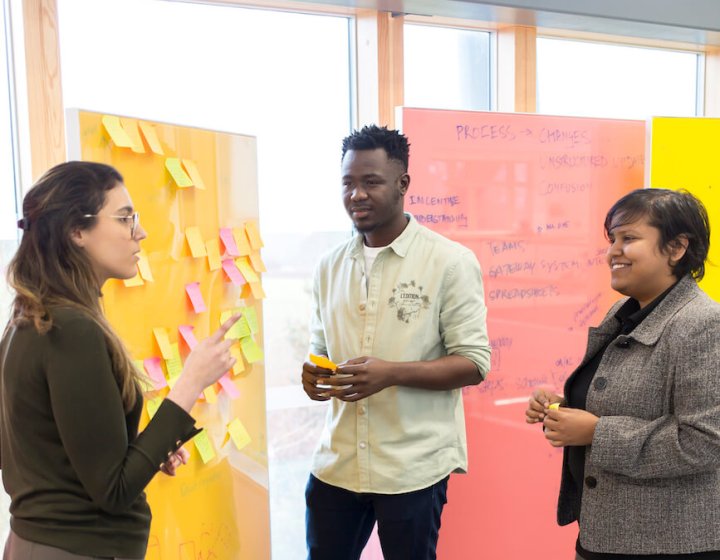 Three people standing in front of a yellow board covered in post-it notes