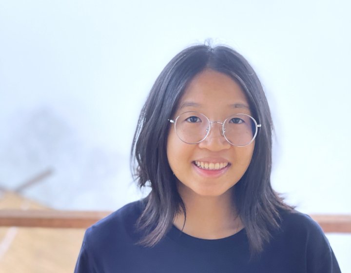 Photo of a student with shoulder length dark hair wearing round gold-framed glasses and a black t-shirt. 