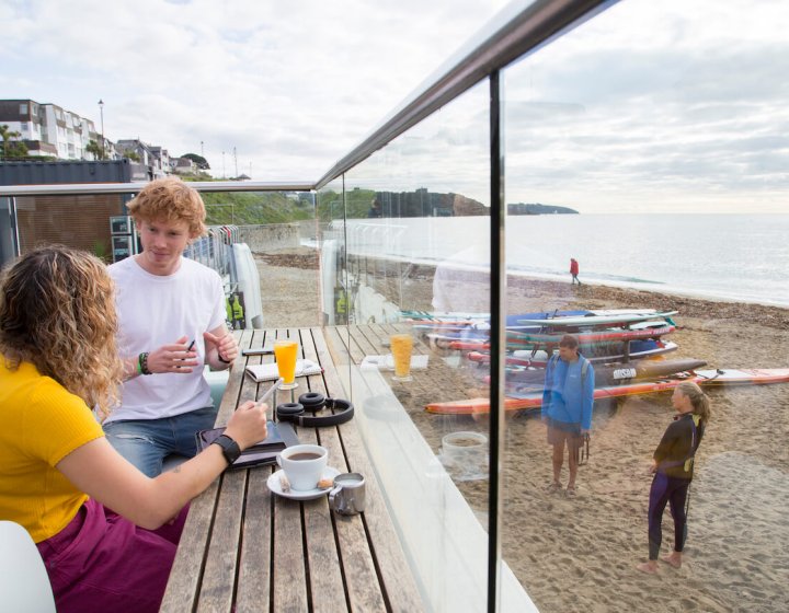 Falmouth University students sat on the terrace at Gylly Beach Cafe with the sea in the background