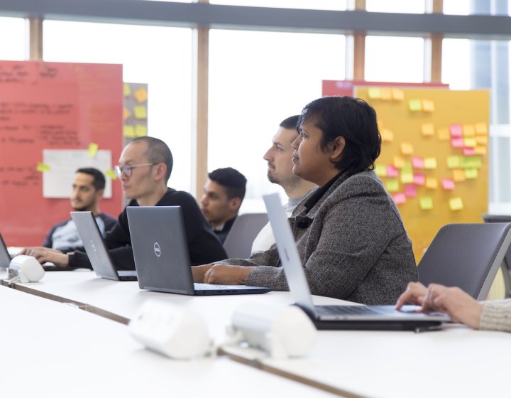 A group of people sat with laptops on a table, board of post-it notes behind them