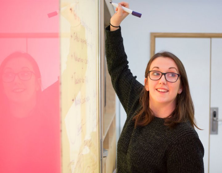 A woman wearing a black jumper, writing on a whiteboard with a pink panel