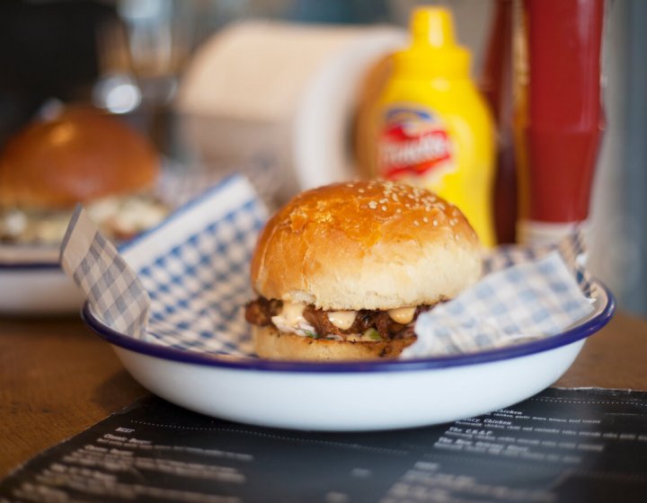 A burger in a white metal bowl with a bottle of mustard in the background