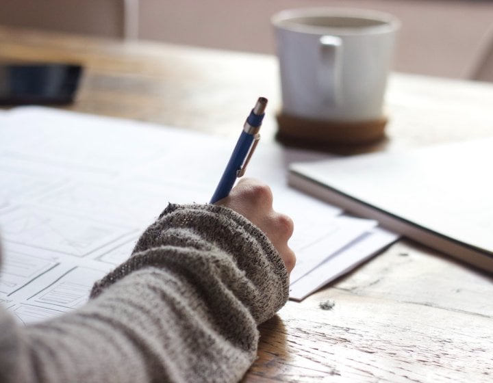 A hand holding a pen writing on paper with a mug in the background