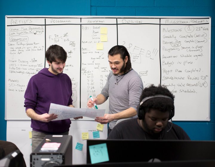 Two students stand in front of a whiteboard scrutinizing a piece of paper. A student sits in the foreground working on a laptop. 