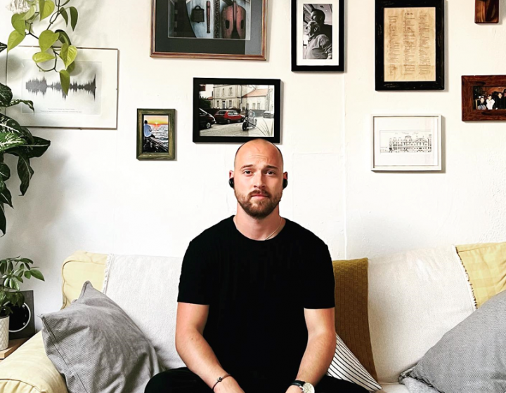 Graduate Thomas Young sitting cross-legged on a sofa with a wall of artwork and photos behind him