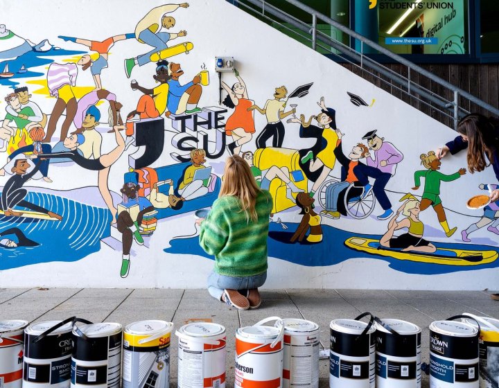 A woman kneels to paint a mural on a wall