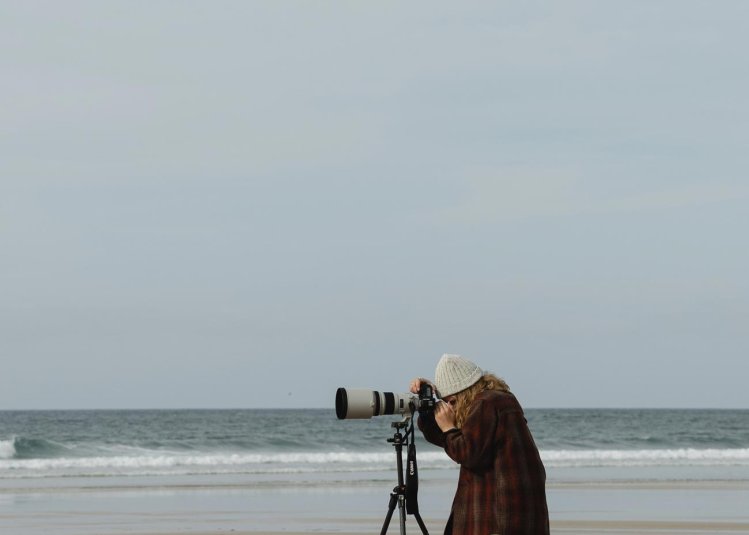 A woman standing on the beach using a large camera on a tripod, 