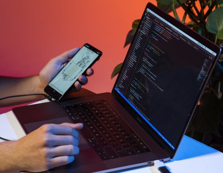 A laptop with code on the screen is connected to a phone with an app open. 