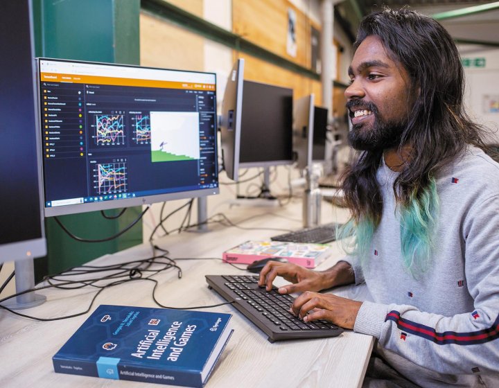 A man typing at a computer with an artificial intelligence for games book on the table