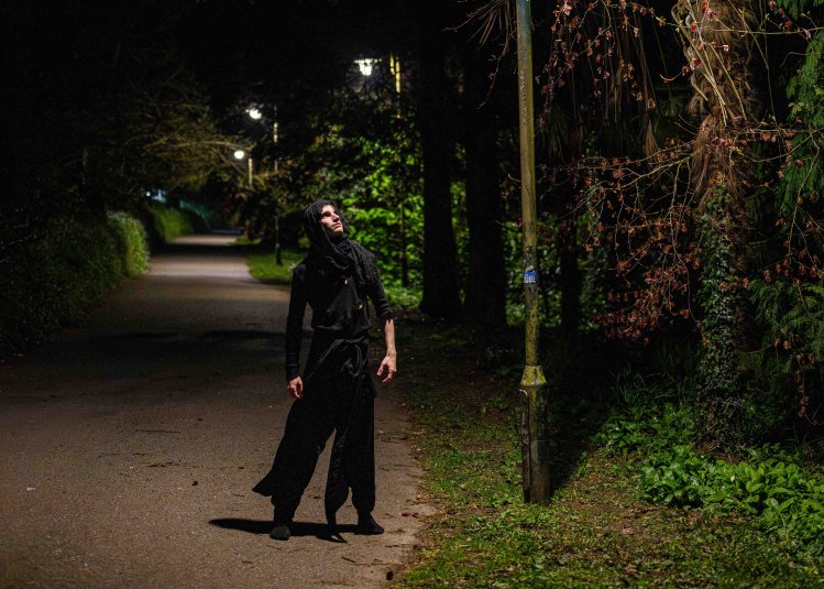 A nighttime image of a person wearing a dark cloak in a road looking up at a streetlight