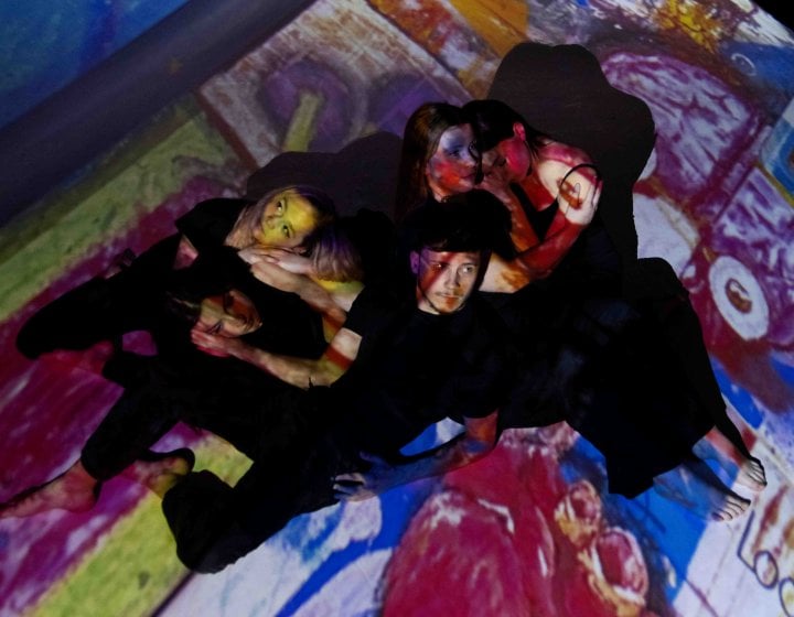 Dancers on stage with a projection of a multi coloured painting overlayed on them